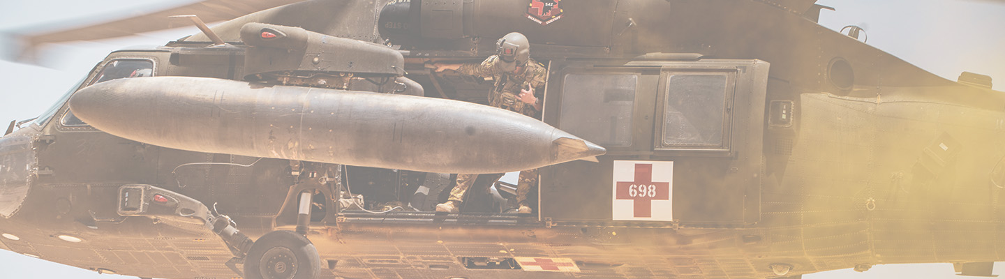 A US Army soldier leaning out of the door of a helicopter as it approaches a landing zone during a training exercise.